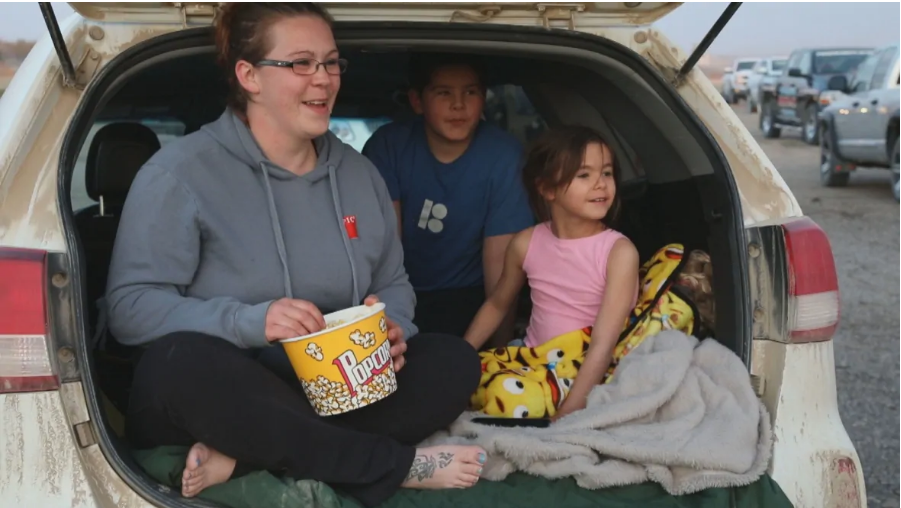 Imagen de una familia compuesta por una madre y dos hermanos, sentados en la parte trasera de su cotche blnaco, riendose por la pelicula que estan viendo.