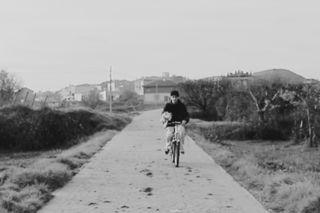 Fotografia en blanc i negre d'un jove circulant en bicicleta per un camí pavimentat enmig d'un entorn natural. Al fons s'observen diverses cases i edificis d'un poble. L'ambient és tranquil i rural, amb vegetació als costats del camí.