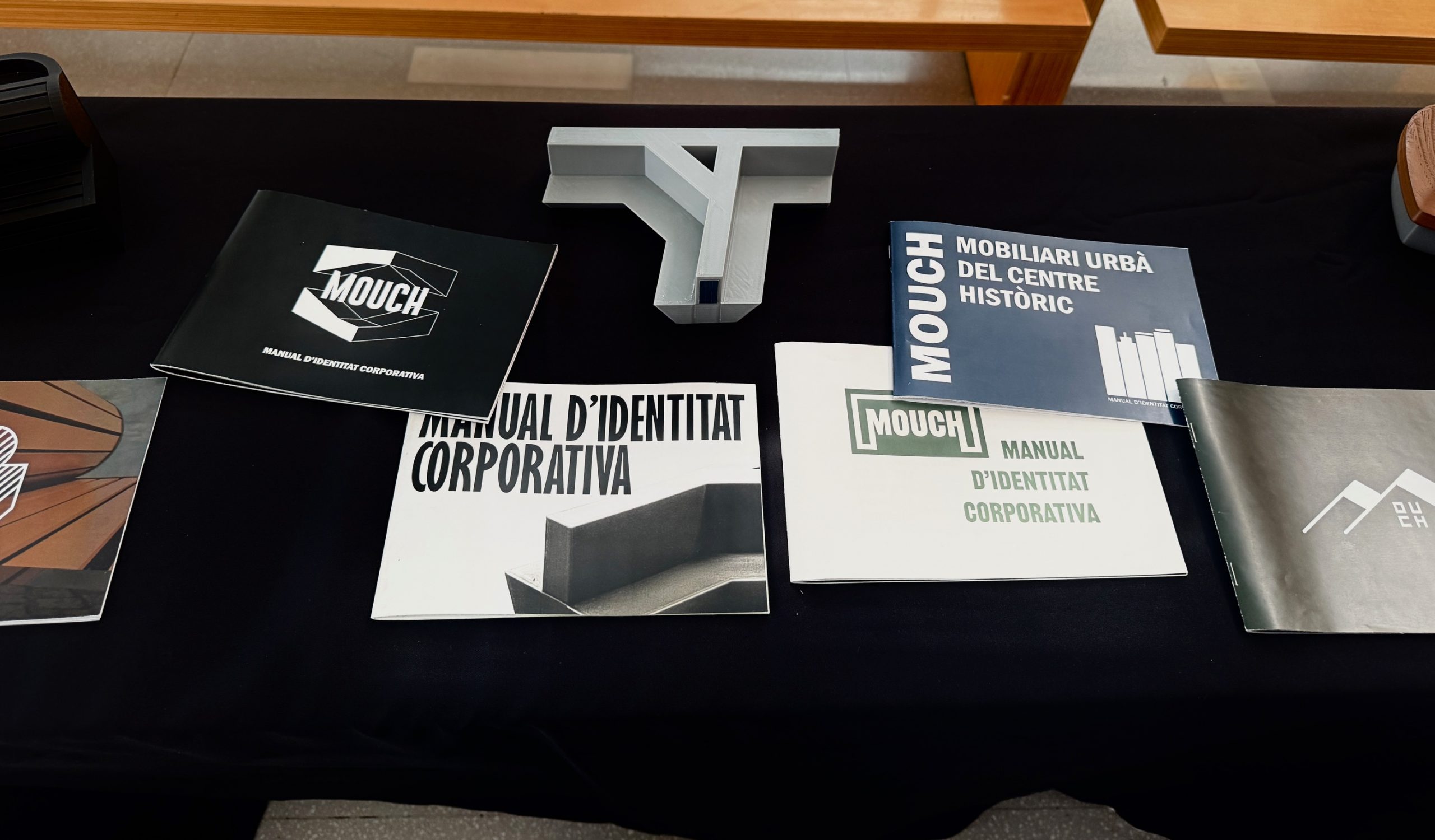 Vista d'una taula amb diversos llibres i manuals de disseny gràfic o identitat corporativa titulats “MOUCH”. Els llibres tenen portades de diferents colors i estils, i al centre hi ha maqueta del banc en escala 1:10, imprès en impressora 3D. La taula és negra i es troba en un entorn interior.