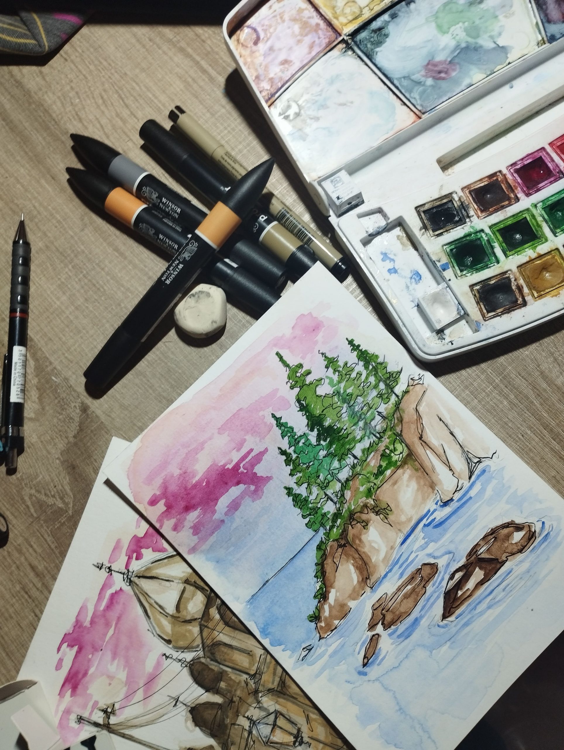 Pintura a l’aquarel·la en un full de paper sobre una taula de fusta. Representa un paisatge amb un pi verd sobre roques marrons al costat d’un llac blau, amb un cel rosa i morat. Al voltant es veuen una caixa d’aquarel·les oberta, diversos retoladors i pinzells negres.