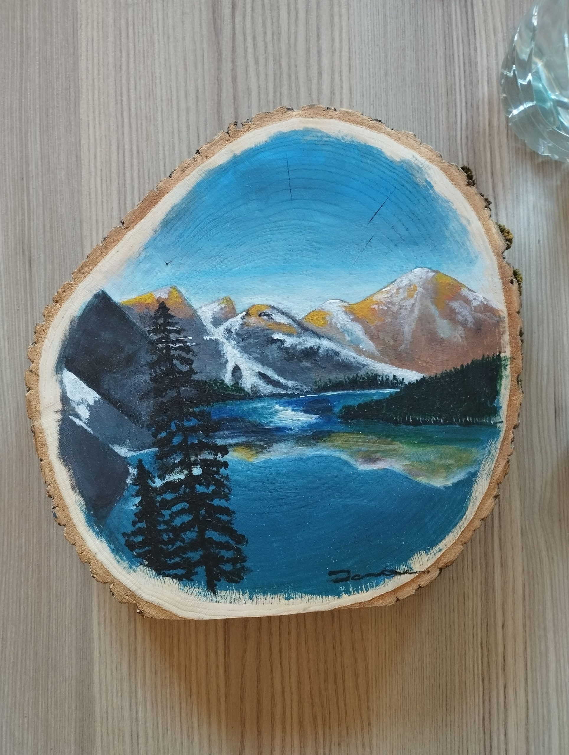 Paisatge pintat a mà sobre una rodella de fusta natural. Es veu un llac blau amb reflexos, flanquejat per pins foscos en primer terme i imponents muntanyes nevades al fons sota un cel blau clar. L’obra està signada a la part inferior dreta.