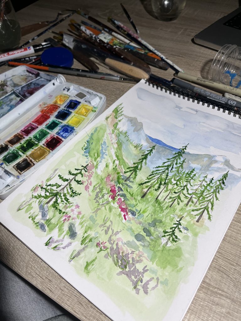 Pintura a l’aquarel·la en un quadern d’espiral oberta sobre una taula. Representa un paisatge de muntanya amb pins verds al primer terme, arbustos amb flors rosades i una muntanya blavosa al fons. Al costat esquerre es veu una caixa d’aquarel·les oberta amb colors vius i diversos pinzells i materials d’art.