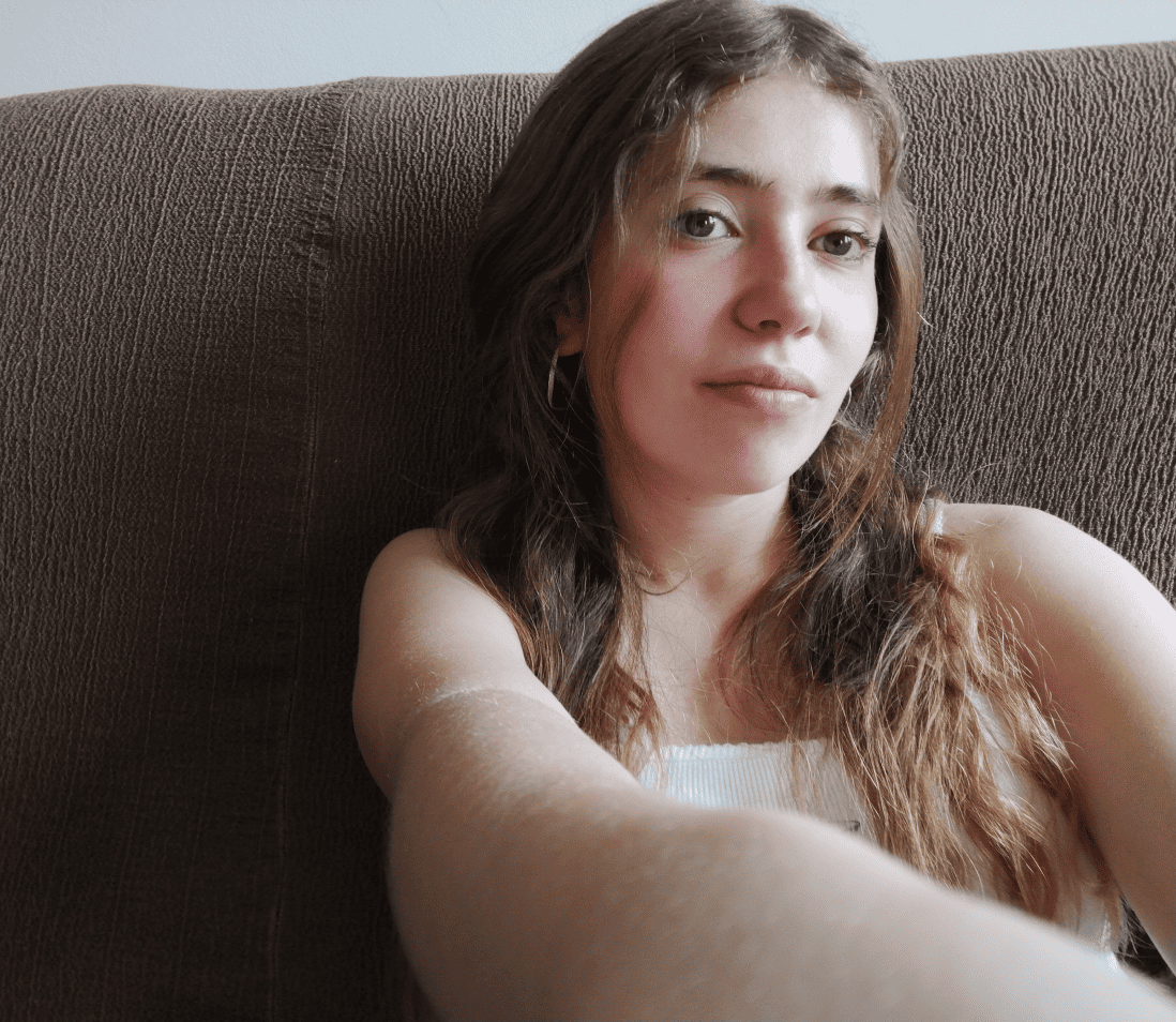 Retrat  de la jana, noia jove amb cabells llargs castanys ondulats, recolzada en un sofà marró. Mira directament a la càmera amb expressió serena i natural, portant una samarreta blanca i arracades. Il·luminació suau i càlida, estil selfie espontani.