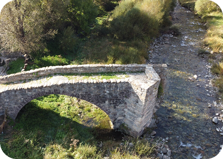 Vista aérea lateral del Puente Viejo de Vilaller, destacando su arco gótico de piedra sobre las aguas claras del río Noguera Ribagorçana. La estructura histórica se muestra rodeada de vegetación típica del Pirineo, reflejando su carácter medieval.