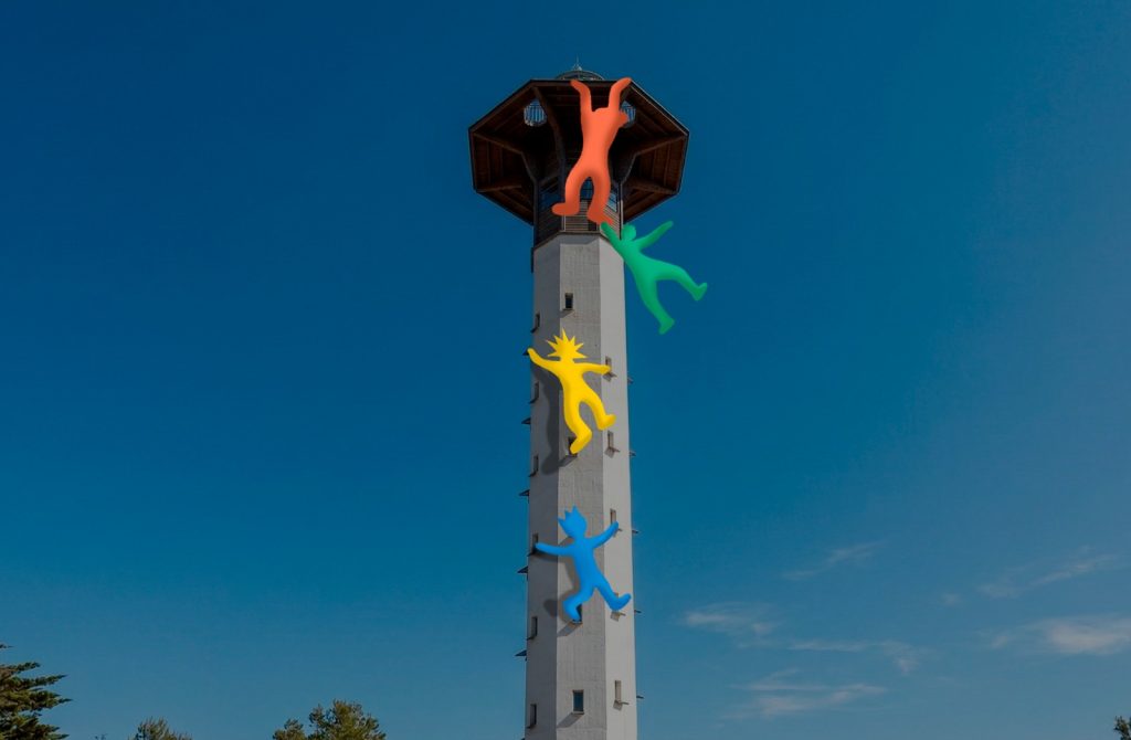 Una fotografía de ángulo contrapicado captura el moderno Faro de Torredembarra, una torre blanca cilíndrica con una plataforma de observación octogonal de madera, bajo un cielo azul despejado. Adosadas al exterior de la torre, cuatro figuras humanas estilizadas y recortadas en colores vibrantes (naranja, verde, amarillo con pelo puntiagudo y azul) están escalando la estructura una encima de la otra, creando una escena dinámica y colorida.
