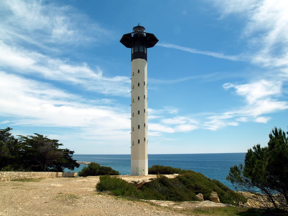 Vista de cuerpo completo del Faro de Torredembarra bajo un cielo azul con nubes dispersas. La estructura destaca por su diseño moderno y esbelto: una torre cilíndrica blanca con pequeñas ventanas rectangulares que culmina en una linterna oscura con una plataforma hexagonal prominente. El faro está situado en un entorno natural rocoso con vegetación mediterránea, frente a la inmensidad del mar azul.
