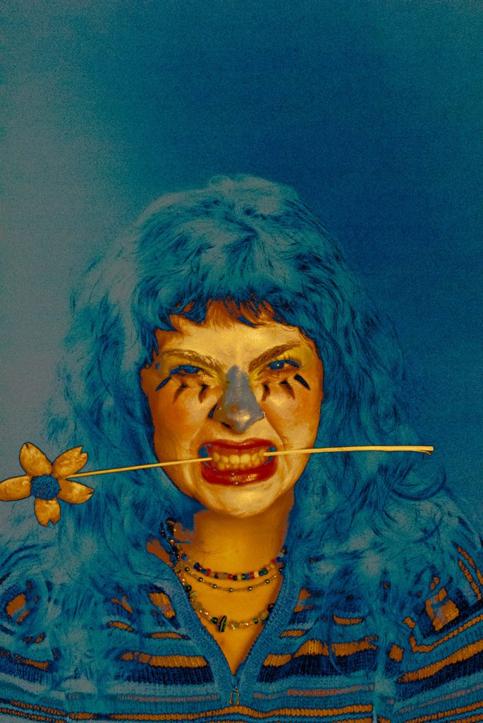 Fotografía artística, se muestra a una mujer con la cara pintada, mordiendo una flor. La mujer, con una expresión furiosa, mira directamente al espectador. Colores invertidos, negativos.